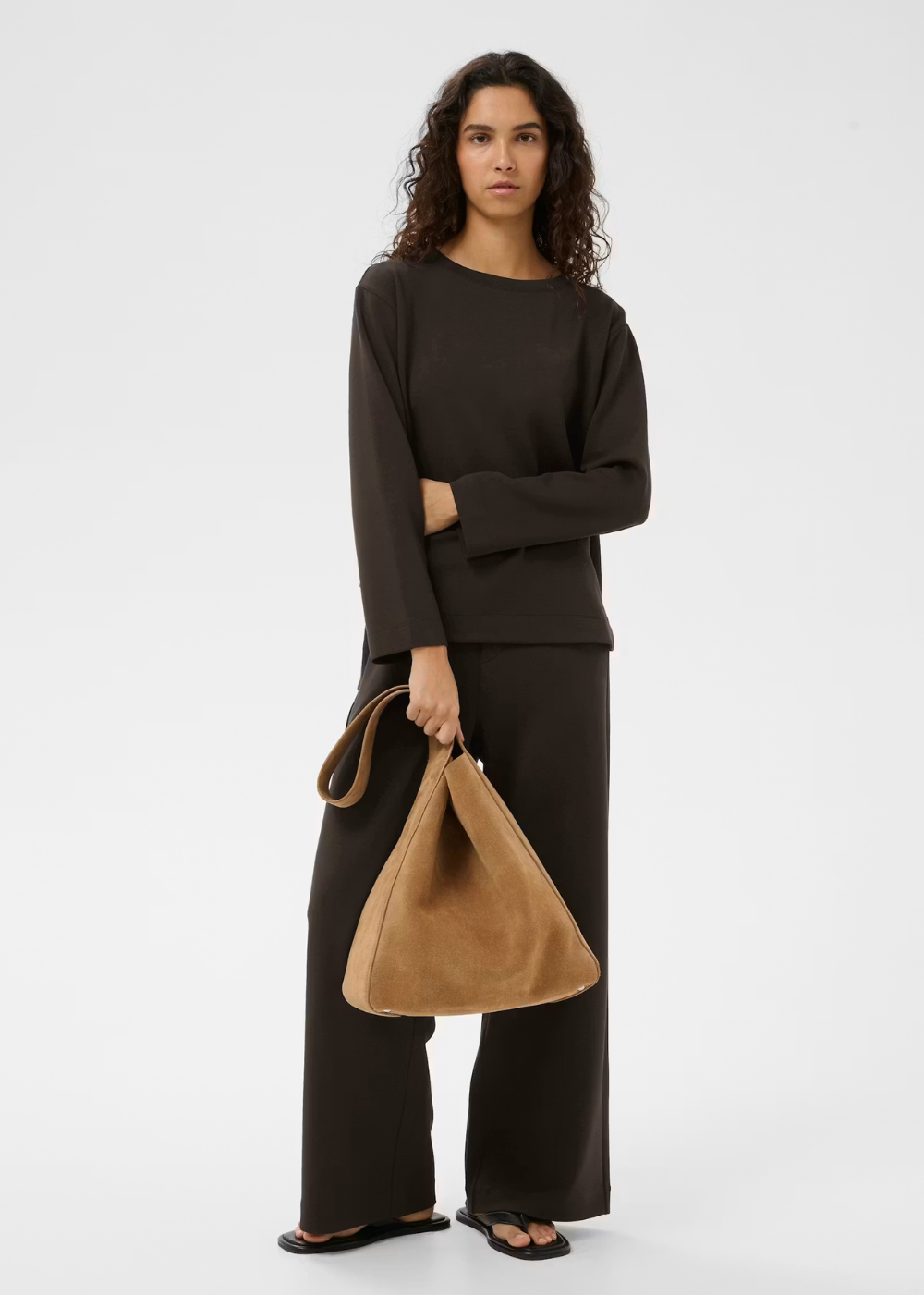 A model stands against a light gray studio background wearing a dark brown long-sleeve top and matching wide-leg pants, holding a tan suede-like shoulder bag.