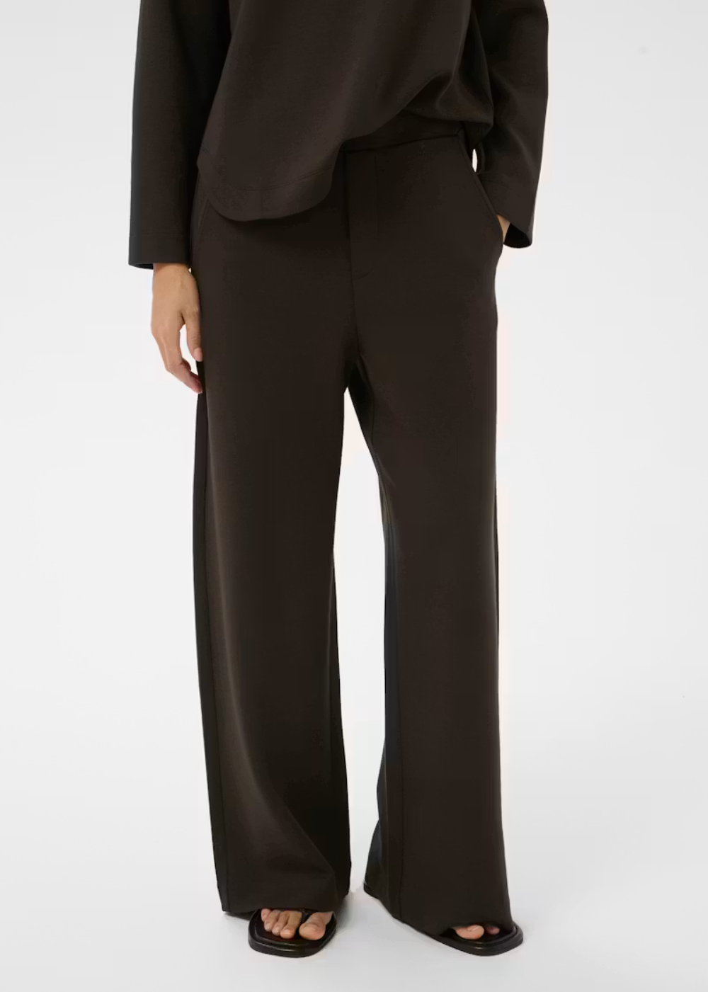 A model wearing dark brown wide-leg trousers with a matching dark brown top and black slide sandals, shown from the waist down against a light gray studio background.