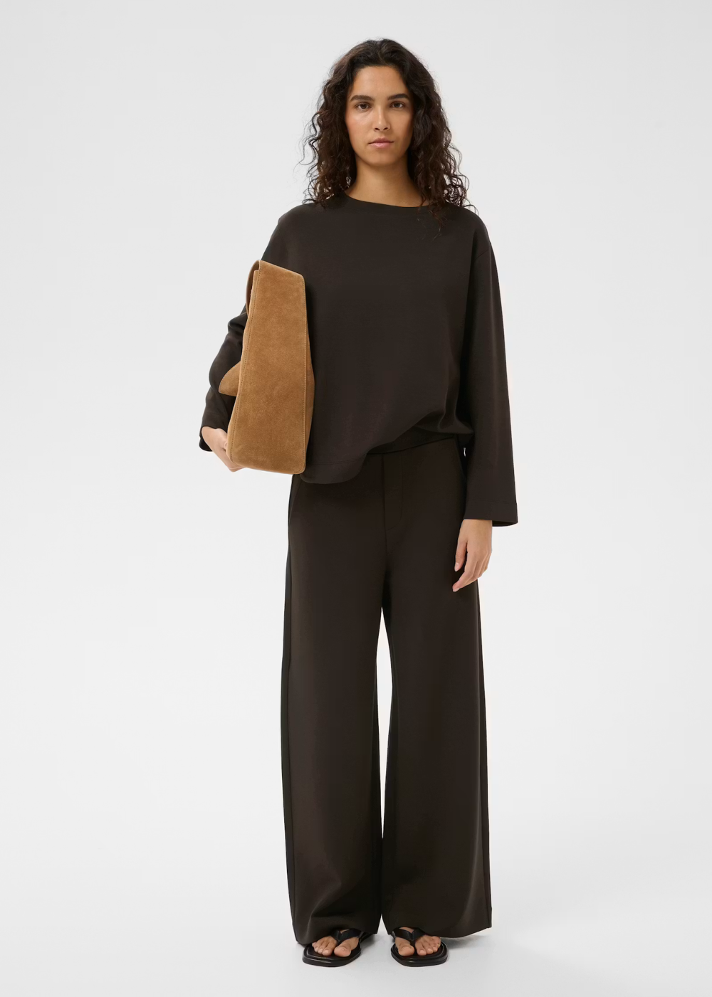 A model stands against a light gray studio background wearing a dark brown wide-leg pants set with a matching long-sleeve top, holding a tan suede-textured clutch.