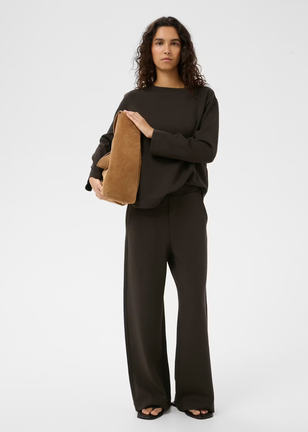 A woman modeling a dark brown long-sleeve top and matching wide-leg trousers, holding a tan suede clutch bag against a plain light gray studio background.