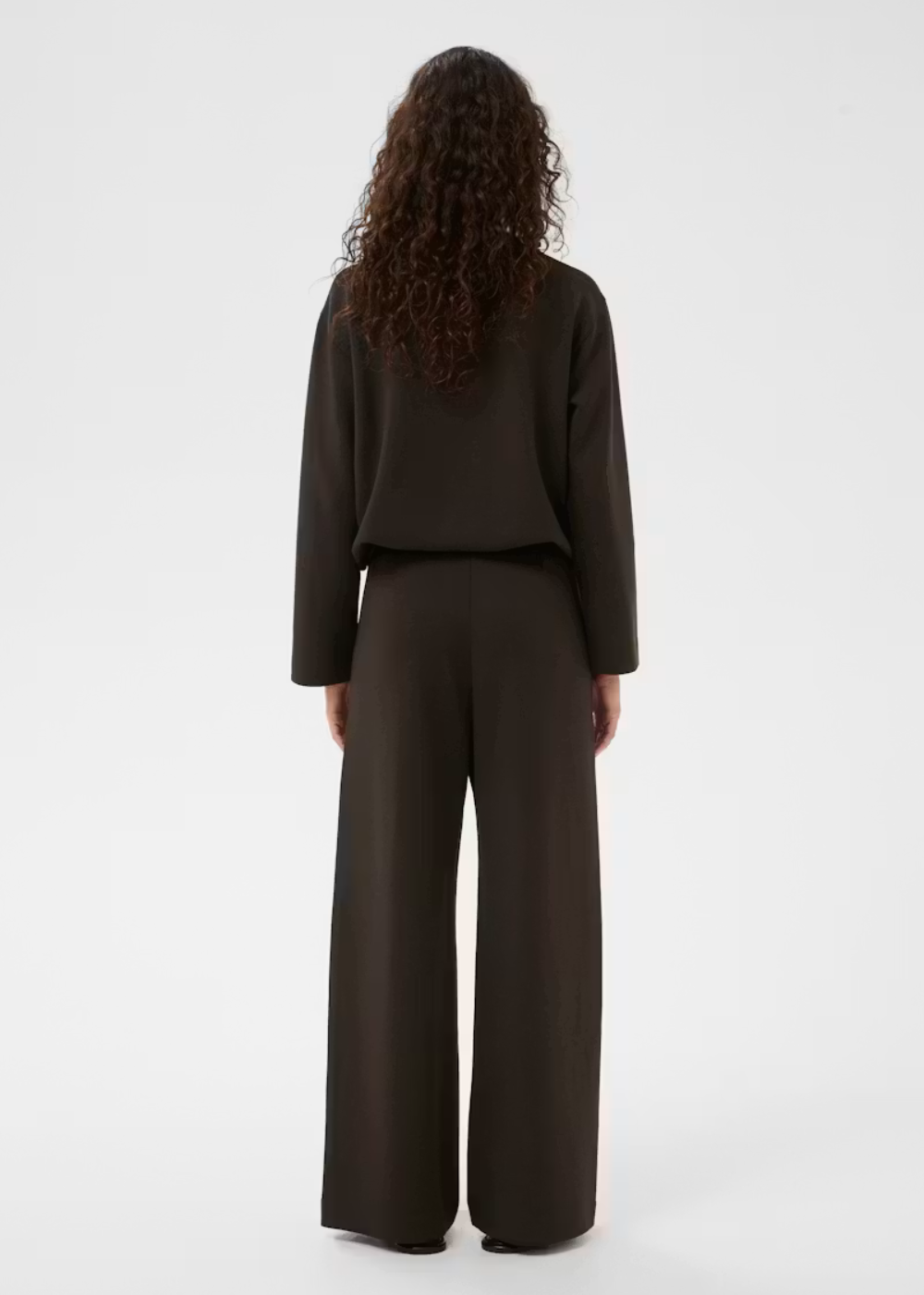 A model viewed from the back wearing a dark brown long-sleeve cropped top and matching wide-leg trousers against a light gray studio background.