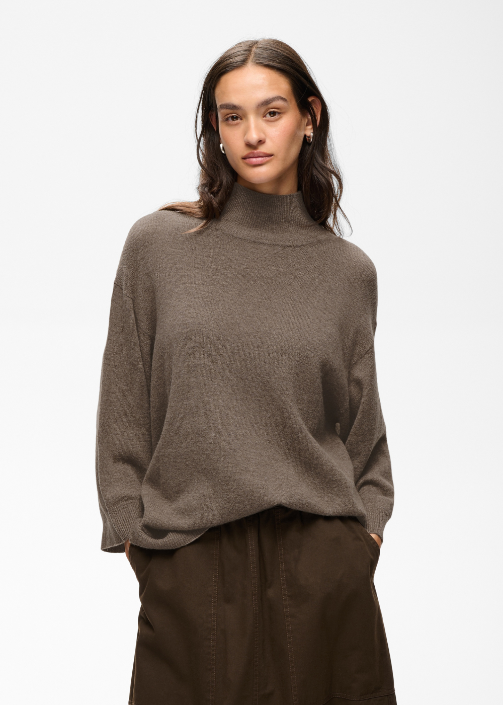 A woman wearing a taupe brown knit mock neck sweater and dark brown trousers, photographed against a clean white studio background.