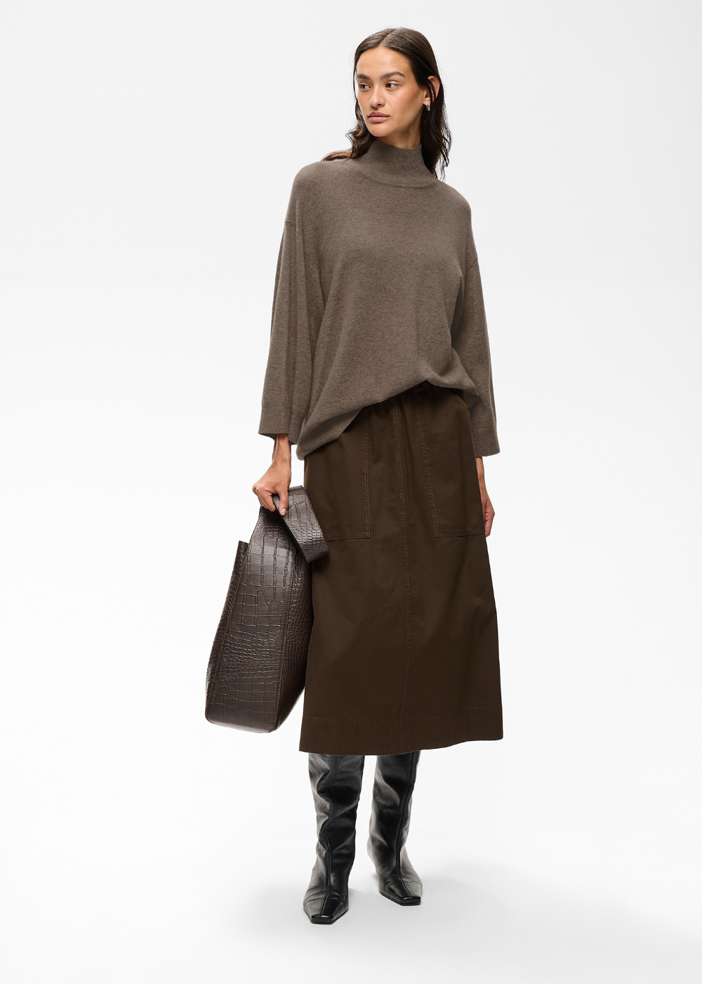 A model stands against a light gray studio background wearing a taupe-brown turtleneck sweater, a dark brown midi skirt, black knee-high boots, and holding a dark brown textured tote bag.