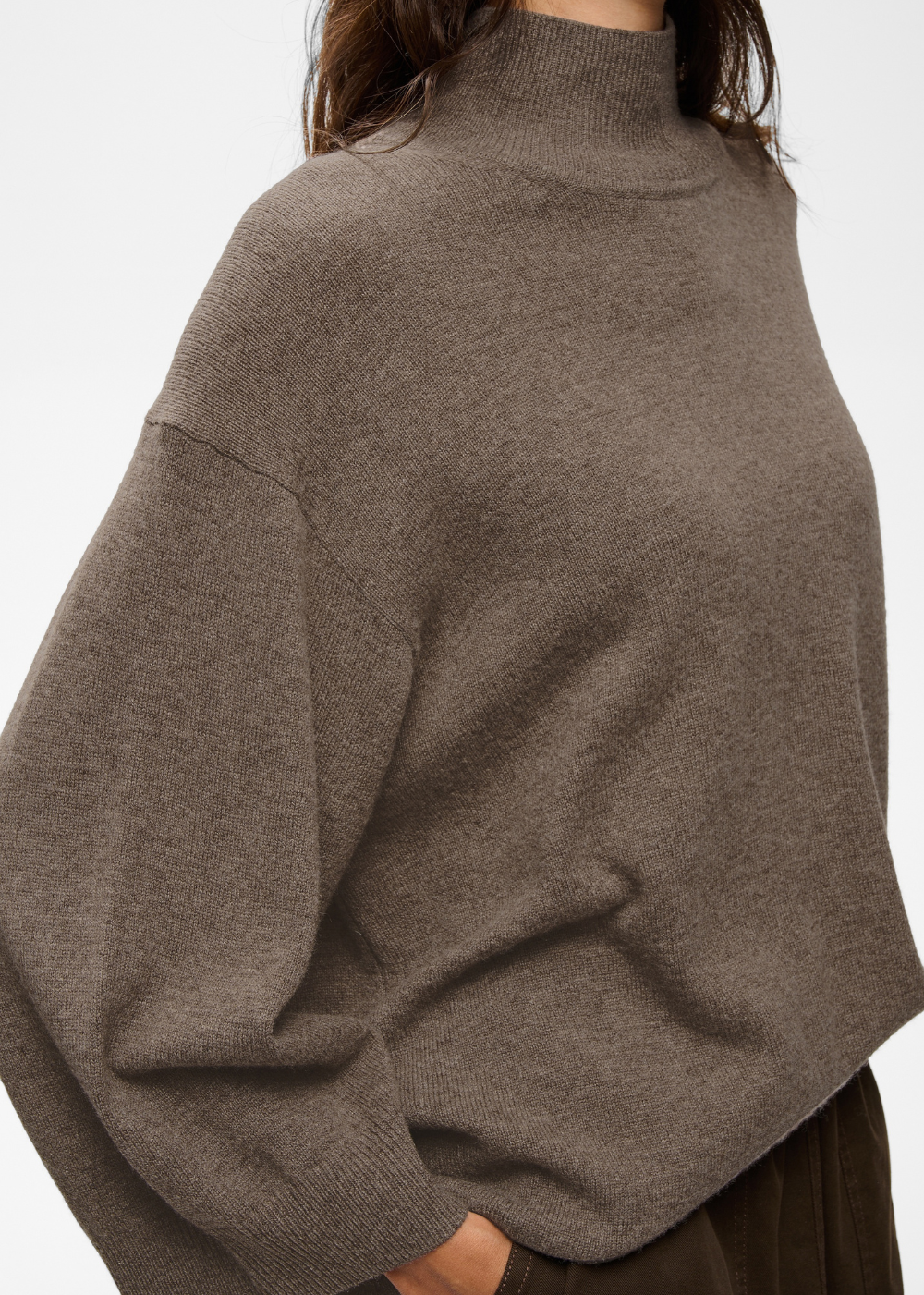 A close-up of a person wearing an oversized taupe-brown knit mock-neck sweater against a plain light background.