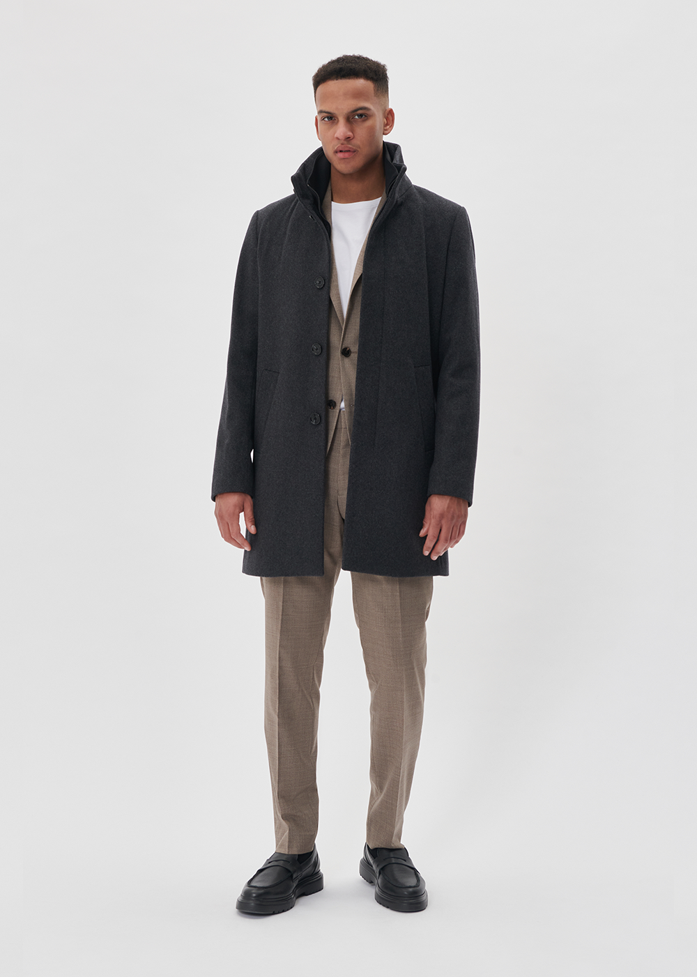 Man wearing a dark grey coat over a beige outfit on a white background