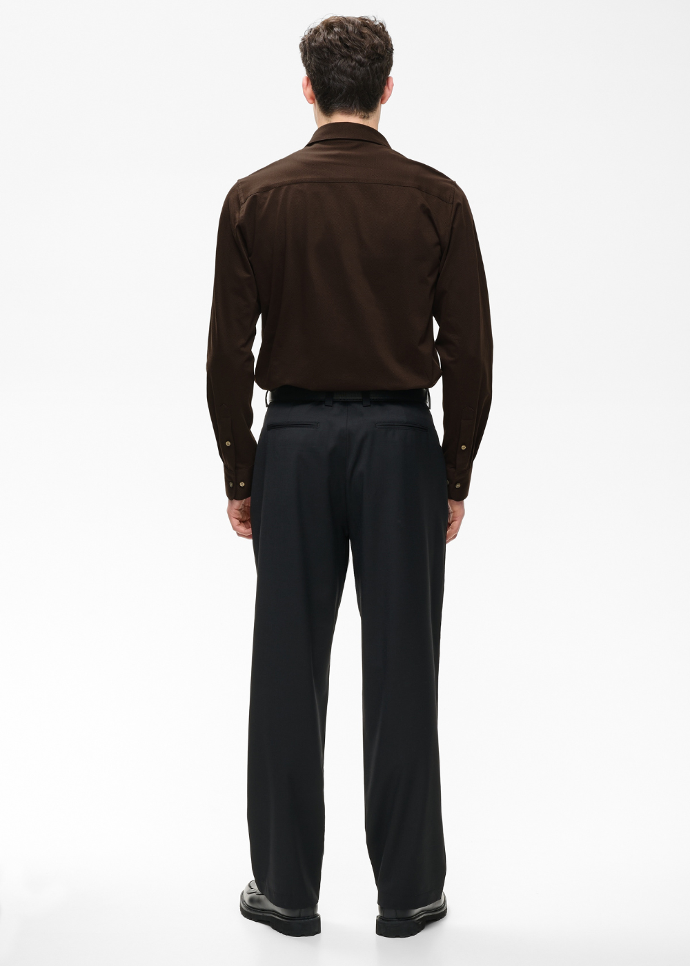 A man viewed from the back wearing a dark brown long-sleeve button-up shirt and black trousers, standing against a plain white studio background.