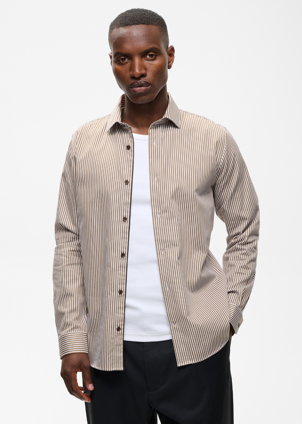 A man wearing a beige and white vertically striped button-down shirt over a white t-shirt and black trousers, photographed against a light gray studio background.