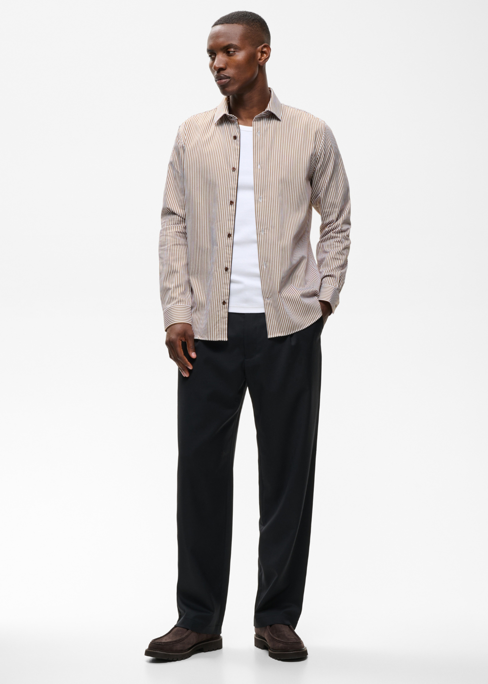 A man modeling a light beige and white vertically striped button-down shirt over a white t-shirt, paired with black trousers and brown shoes, against a plain white studio background.