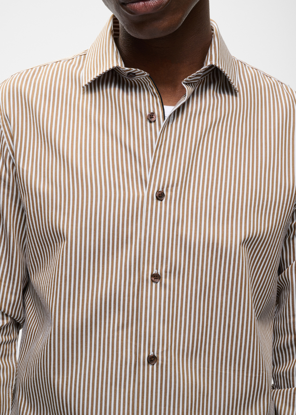 A close-up of a man wearing a beige and brown vertically striped button-down shirt, showing the collar, front buttons, and part of the chest against a plain light background.