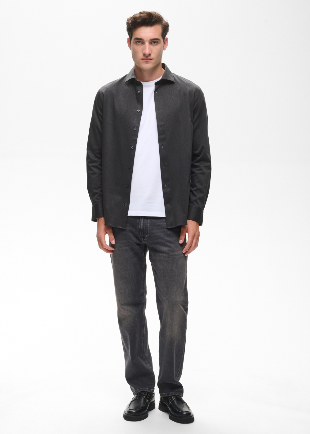 A man modeling a black long-sleeve button-up shirt over a white t-shirt, paired with dark gray jeans and black shoes, against a plain white studio background.