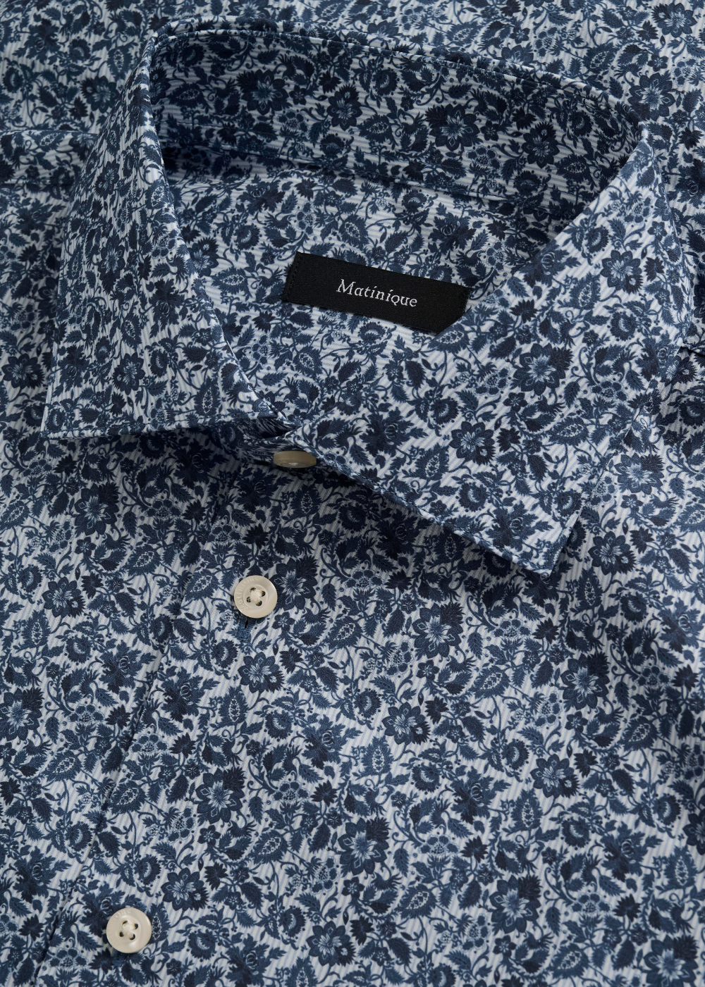 Close-up of a collared button-up shirt with a dense navy and white floral print, featuring a black neck label that reads 'Matinique'.