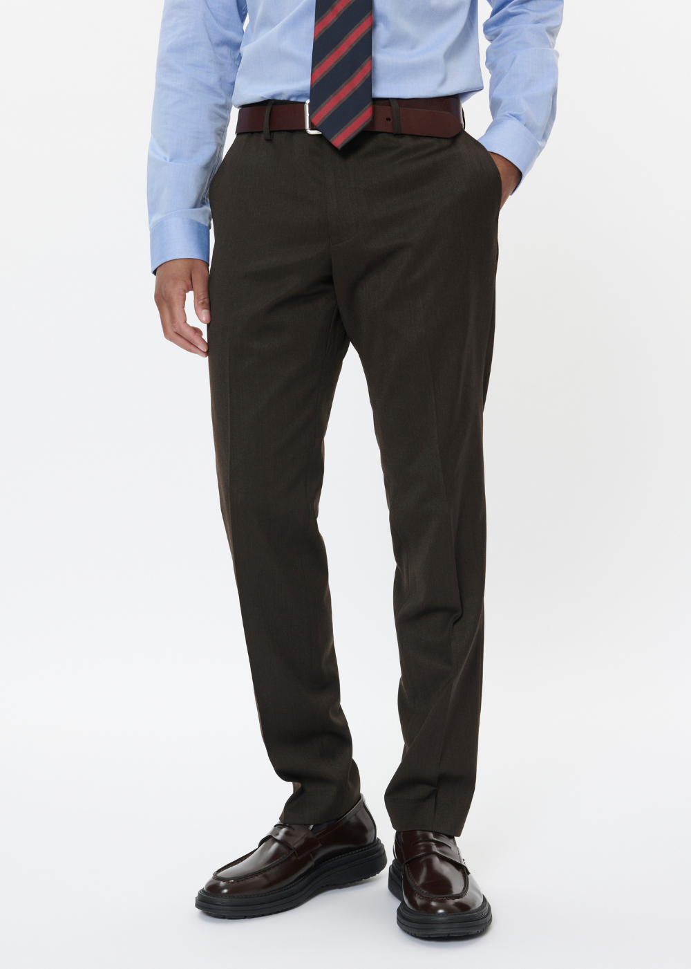A man wearing dark brown dress pants, a light blue dress shirt, a striped tie, a brown belt, and dark brown leather dress shoes, shown from the waist down on a white studio background.
