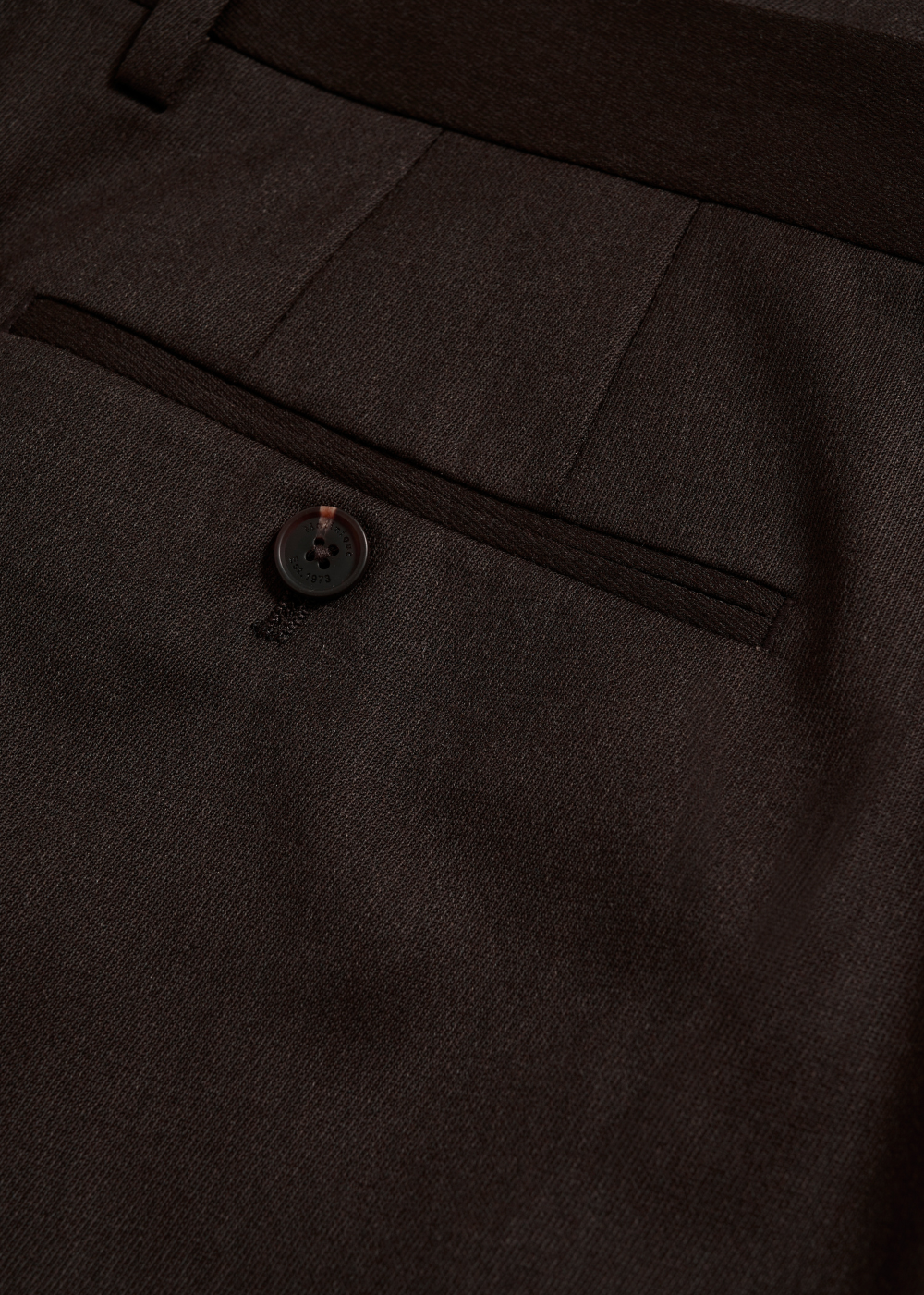 Close-up of dark brown tailored trousers showing a front welt pocket with a single button closure and the fine fabric texture.
