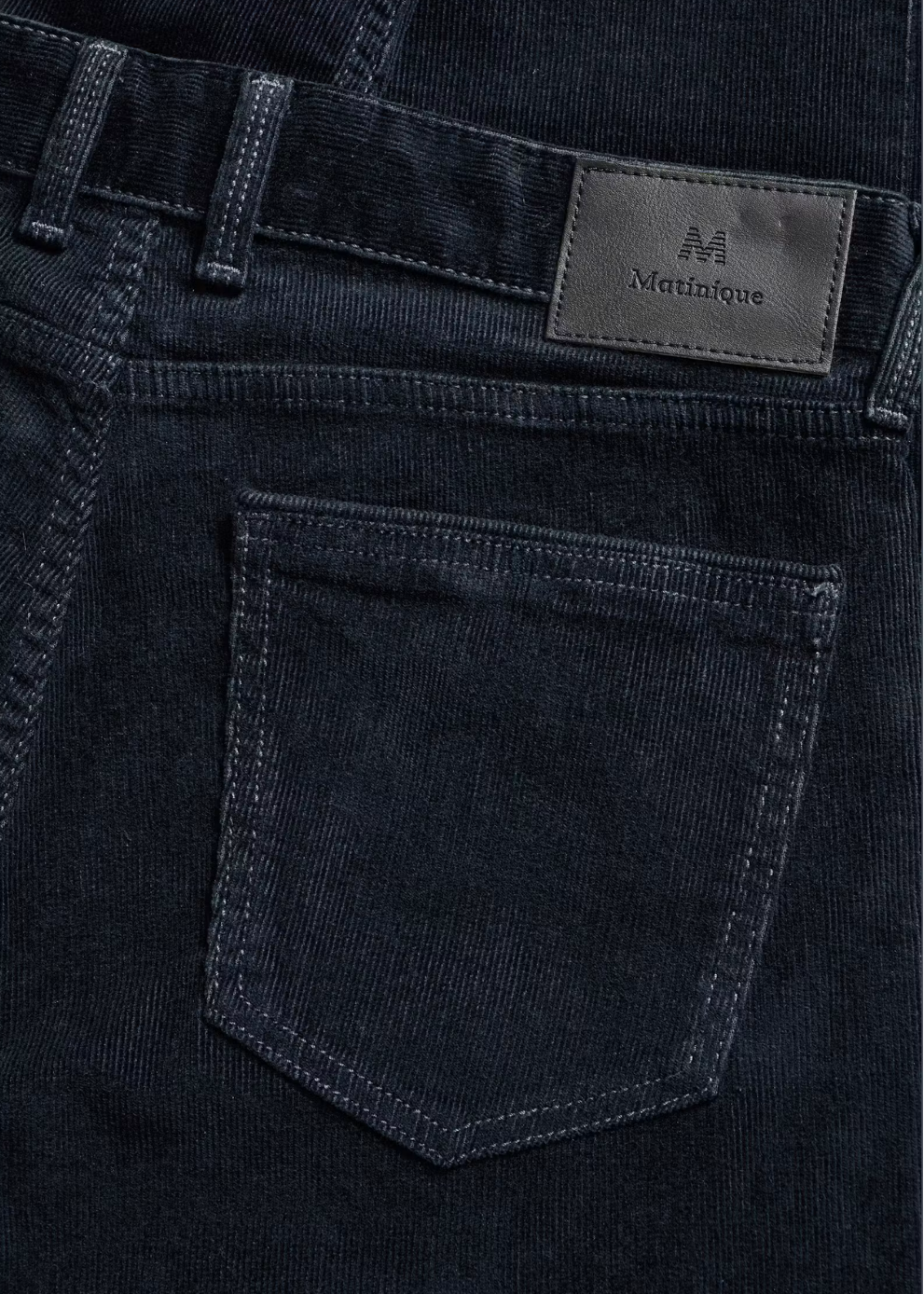 Close-up of the back waistband and pocket area of dark indigo corduroy jeans, showing a black leather patch with the Mattinique brand logo.