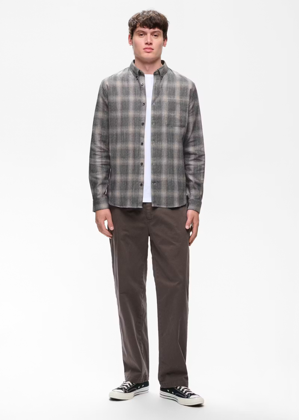 A man modeling a gray and black plaid button-down shirt over a white undershirt, paired with dark brown pants and black sneakers, against a plain white studio background.