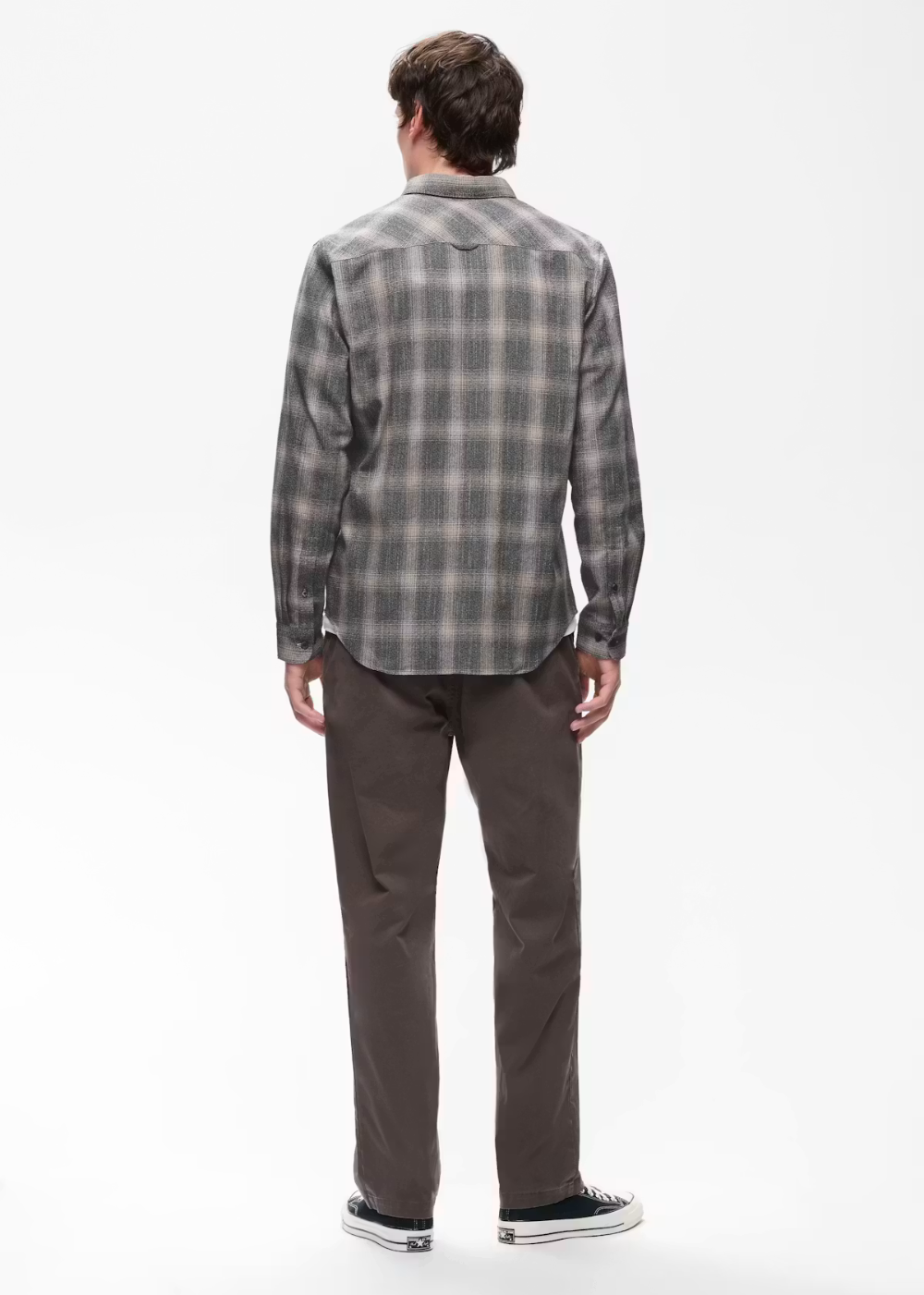 A man viewed from behind wearing a gray and black plaid button-down shirt, dark brown pants, and black sneakers with white soles, set against a plain white studio background.