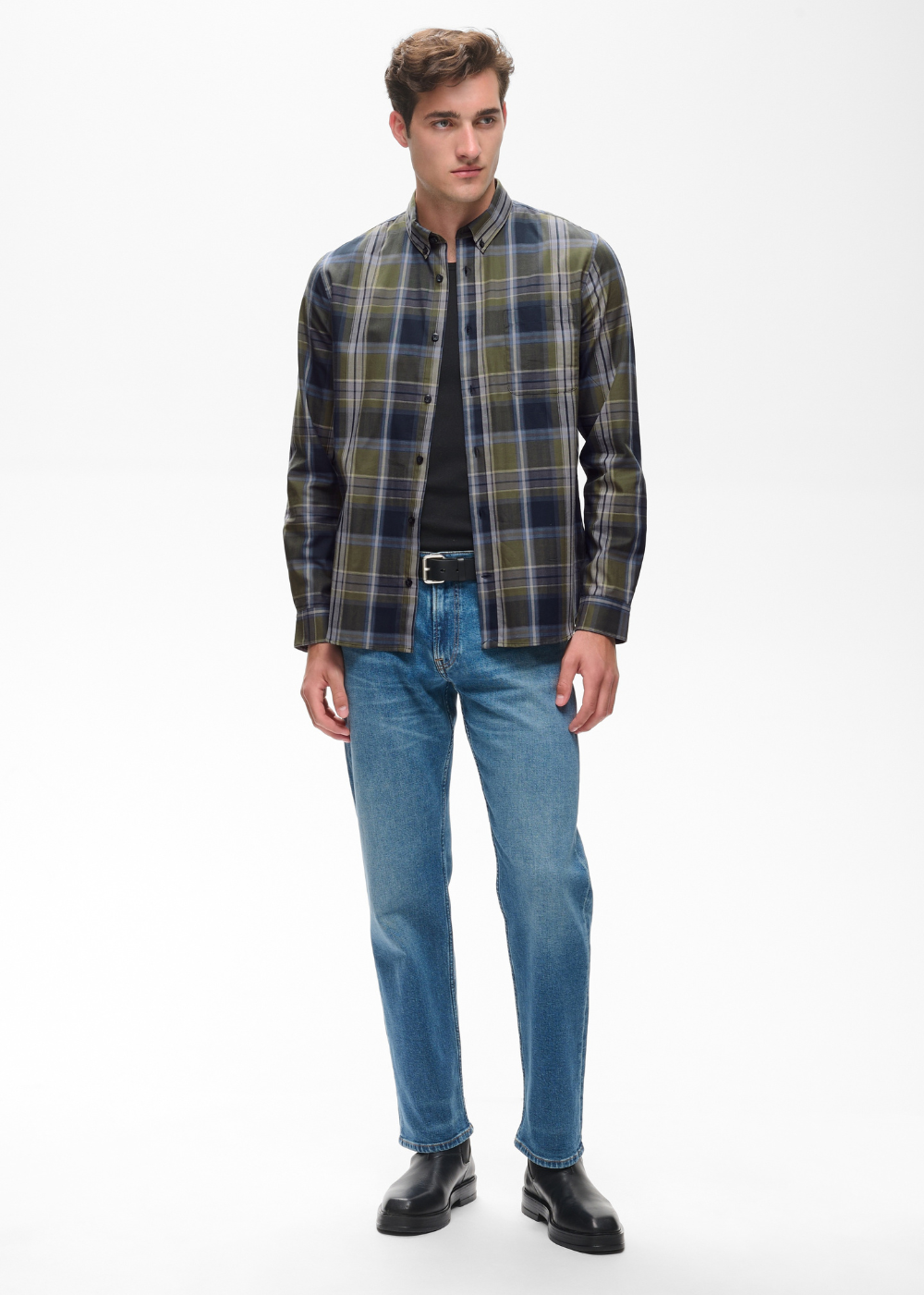 A man modeling a dark green and navy plaid overshirt over a black shirt, paired with blue jeans and black leather shoes, against a plain white studio background.