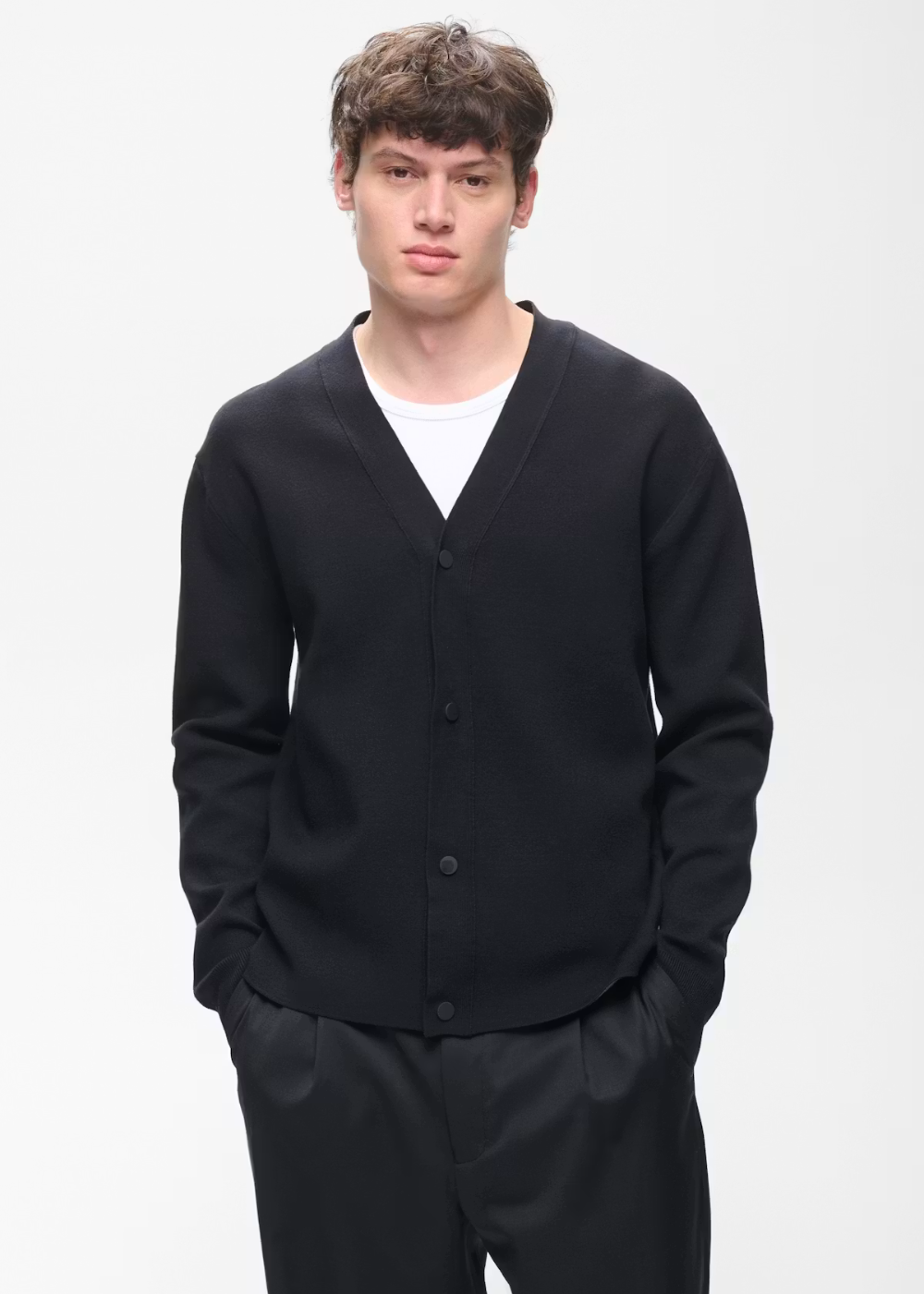 A young man modeling a black button-up knit cardigan over a white t-shirt and black trousers, photographed against a clean white studio background.