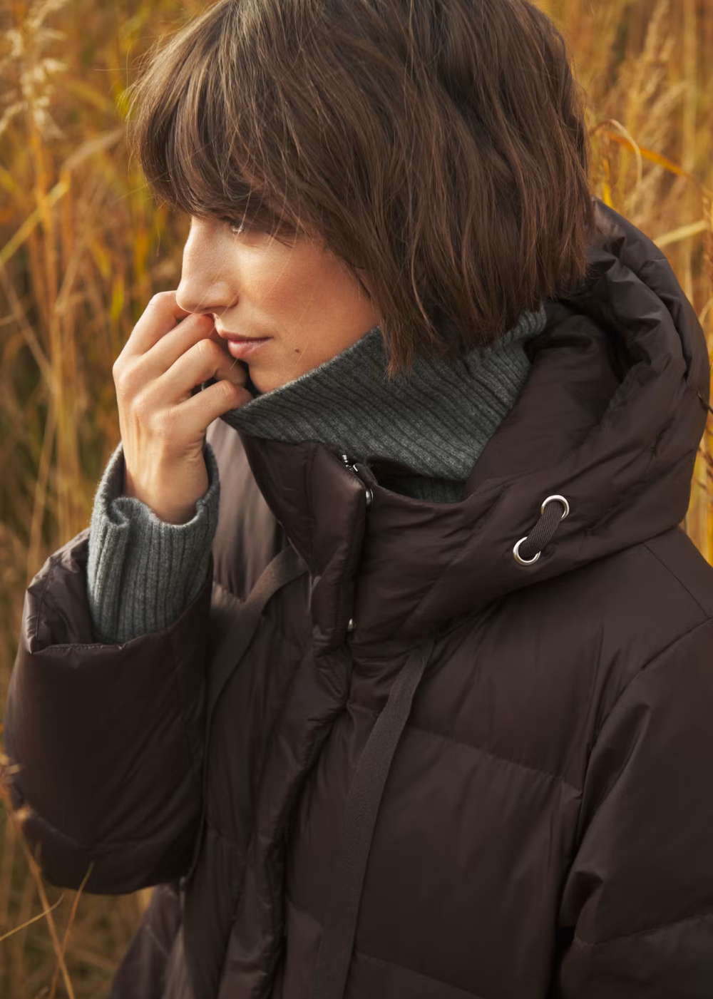 A woman wearing a dark brown puffer jacket over a gray ribbed turtleneck sweater, standing outdoors in a field of dry golden grass.