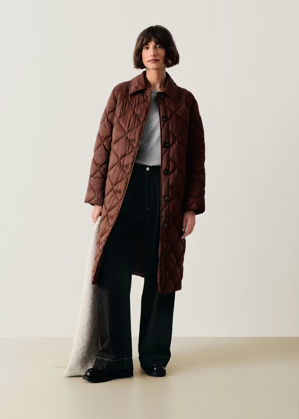 A model stands against a plain light beige studio background wearing a long brown quilted coat over a gray top and black wide-leg pants.