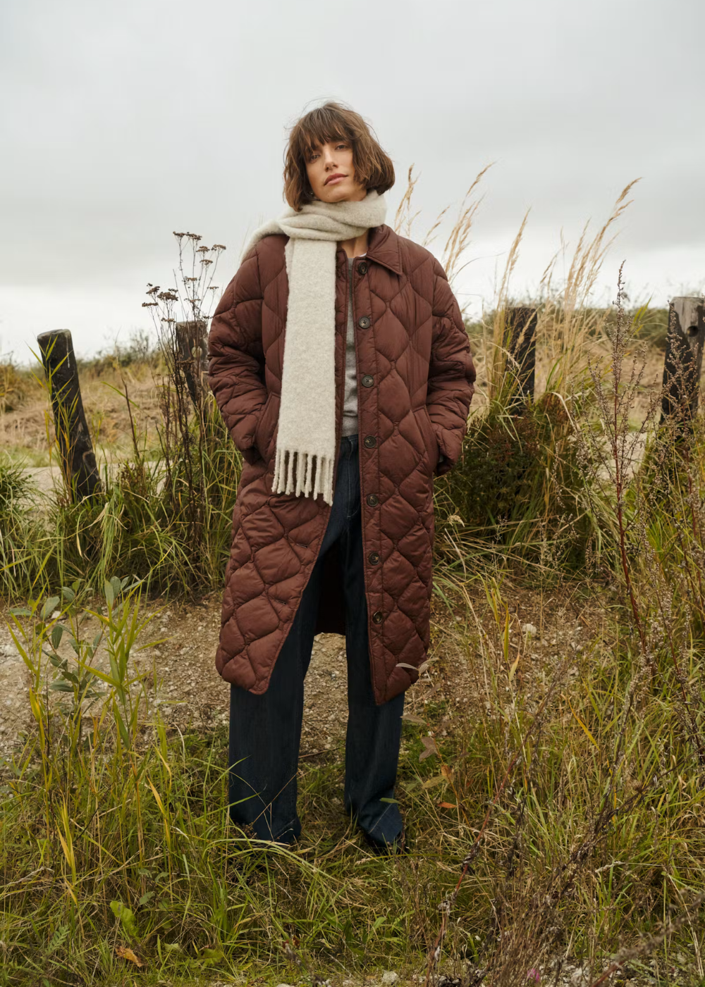 A model stands outdoors in a grassy field under an overcast sky, wearing a long brown quilted coat, a cream knit scarf, and dark wide-leg trousers.