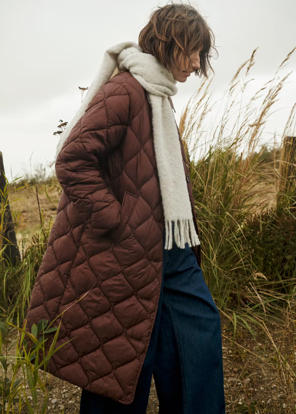 A model wearing a brown diamond-quilted puffer coat, a thick white knit scarf, and blue jeans, standing in a grassy outdoor field under an overcast sky.