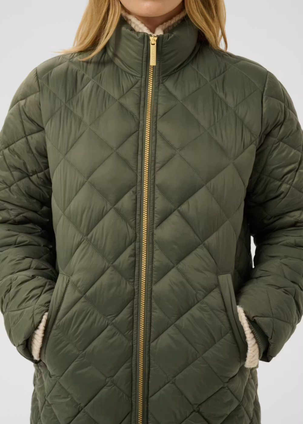 An olive green diamond-quilted puffer jacket with a gold-tone front zipper and a cream-colored fleece lining, shown on a model against a light gray background.