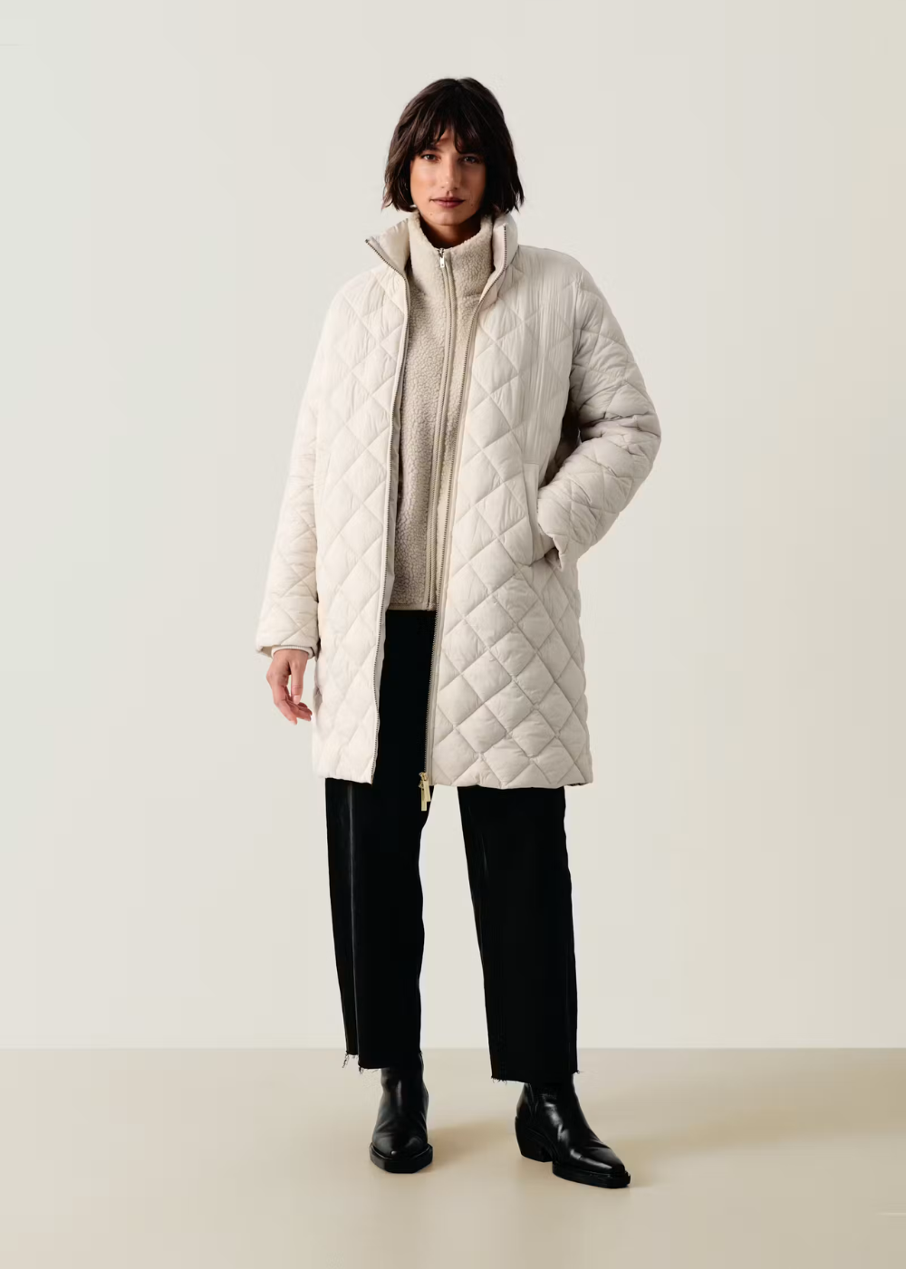 A model stands against a plain light beige studio background wearing a long quilted puffer coat in off-white, a cream ribbed turtleneck sweater, black wide-leg trousers, and black leather boots.