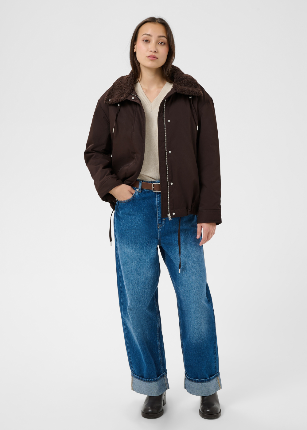 A model stands against a light gray studio background wearing a dark brown hooded jacket over a cream top, blue wide-leg jeans, and black boots.