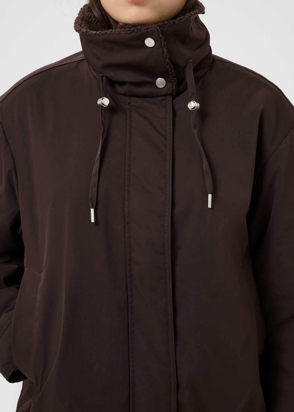 A close-up of a dark brown hooded jacket with a high collar, silver snap buttons, and drawstrings, worn by a model against a light gray background.
