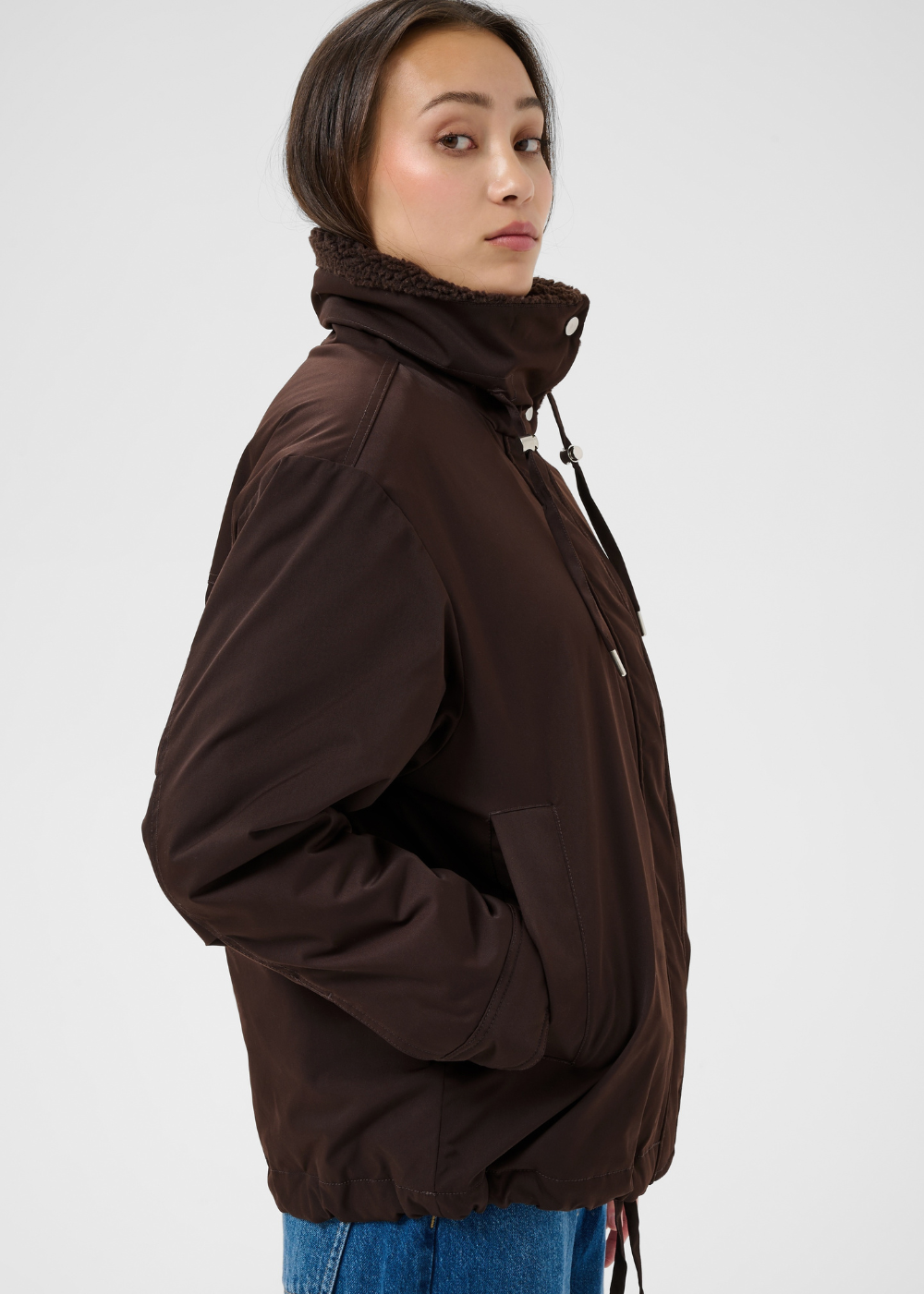 A woman wearing a dark brown lightweight jacket with a high collar and drawstrings, paired with blue jeans, photographed against a light gray studio background.