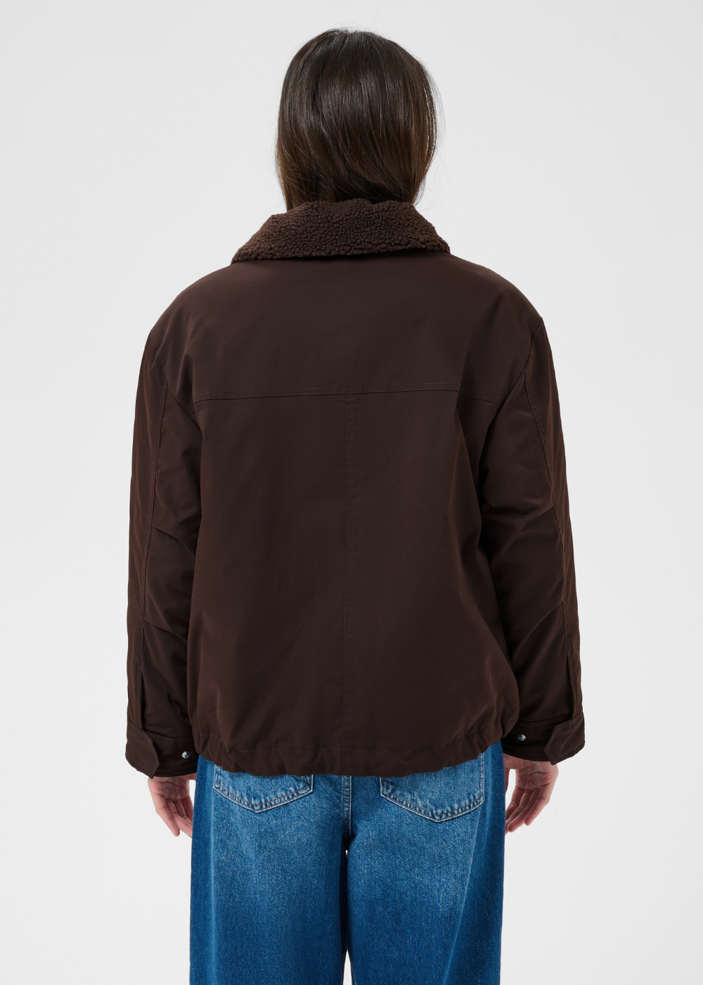 A person shown from the back wearing a dark brown jacket with a fuzzy collar and blue jeans, photographed against a plain light gray studio background.