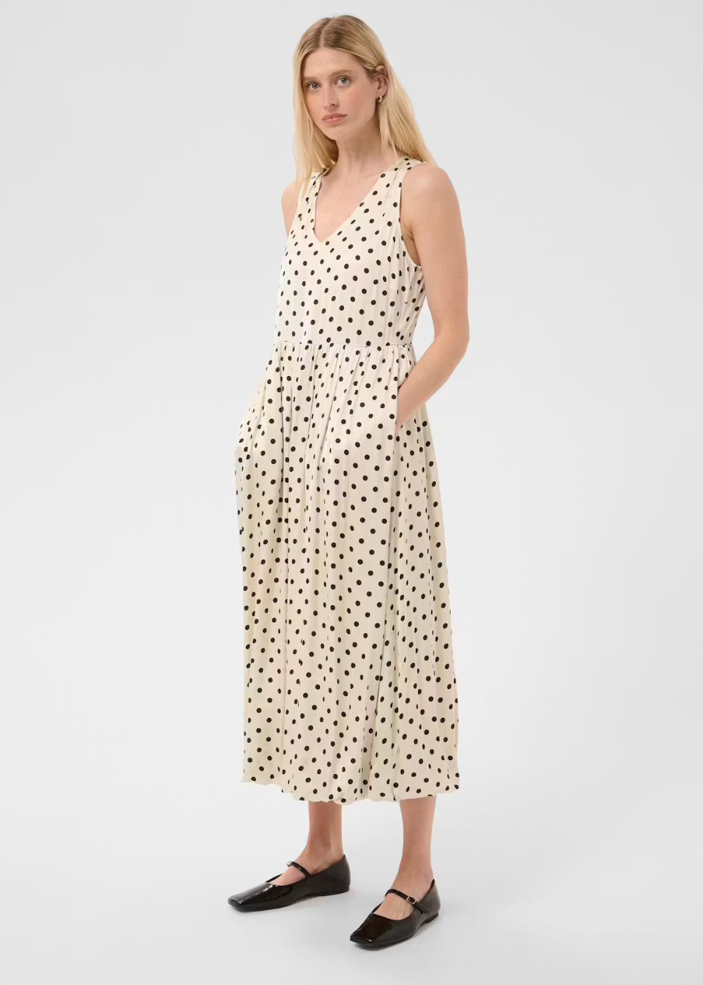 A woman modeling a sleeveless cream midi dress with a black polka dot pattern, featuring a V-neckline, side pockets, and a tie-waist detail, paired with black flat shoes against a light gray studio background.