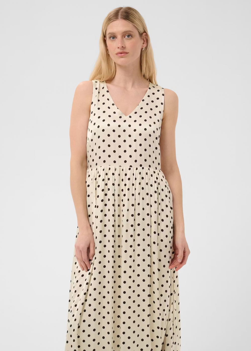A woman modeling a sleeveless V-neck maxi dress with a black polka dot pattern on a cream background, set against a plain light gray studio backdrop.