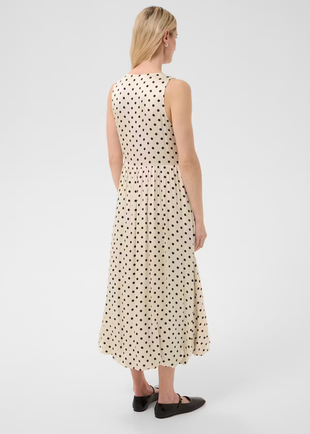 A woman viewed from the back wearing a sleeveless midi dress with a black polka dot pattern on a cream background, paired with black flats, against a light gray studio background.