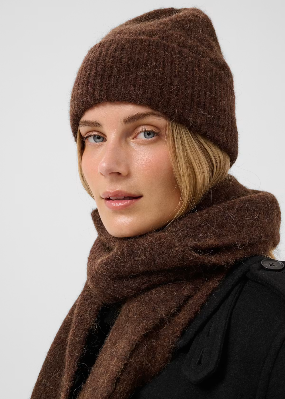 A woman wearing a brown ribbed knit beanie and a matching brown fuzzy scarf, photographed against a light gray studio background.