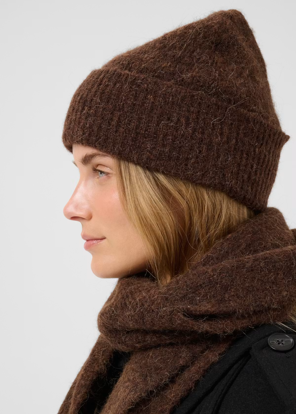 A woman in profile wearing a fuzzy brown knit beanie and matching brown scarf against a light gray studio background.