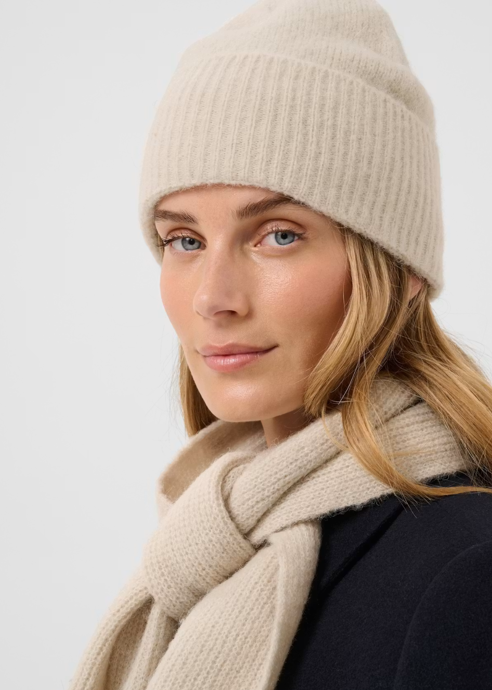 A woman wearing a beige ribbed knit beanie and a matching beige scarf, photographed against a clean light gray studio background.