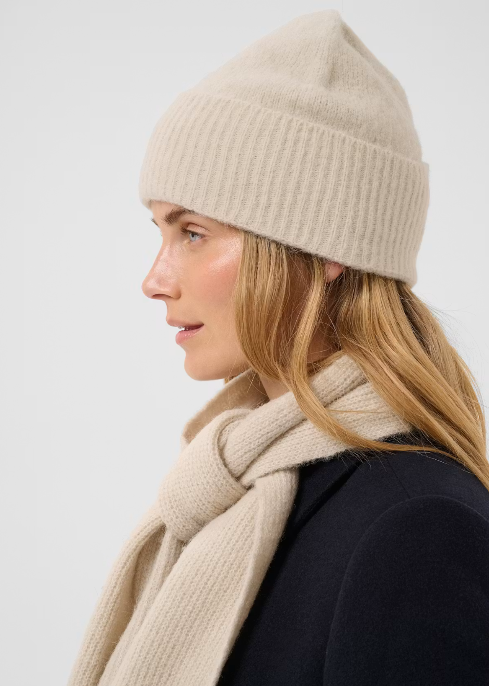 A woman in profile wearing a ribbed beige knit beanie and a matching beige scarf, paired with a dark navy coat, against a light gray studio background.