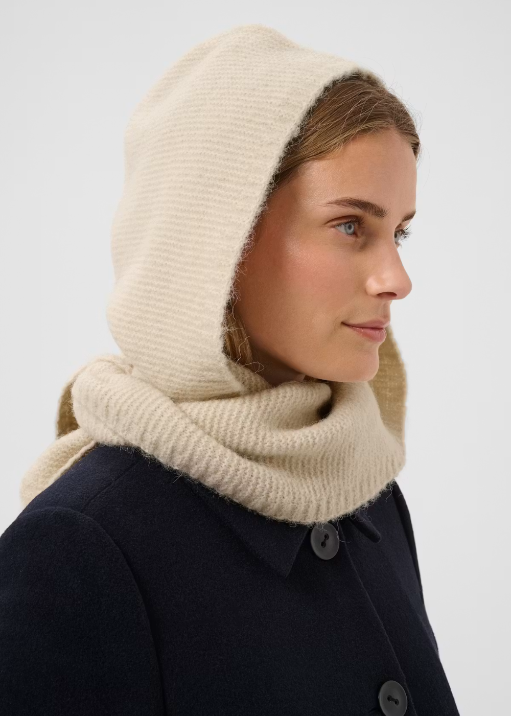 A woman in profile wearing a cream-colored knit hooded cowl neck warmer over a dark navy coat, against a light gray studio background.