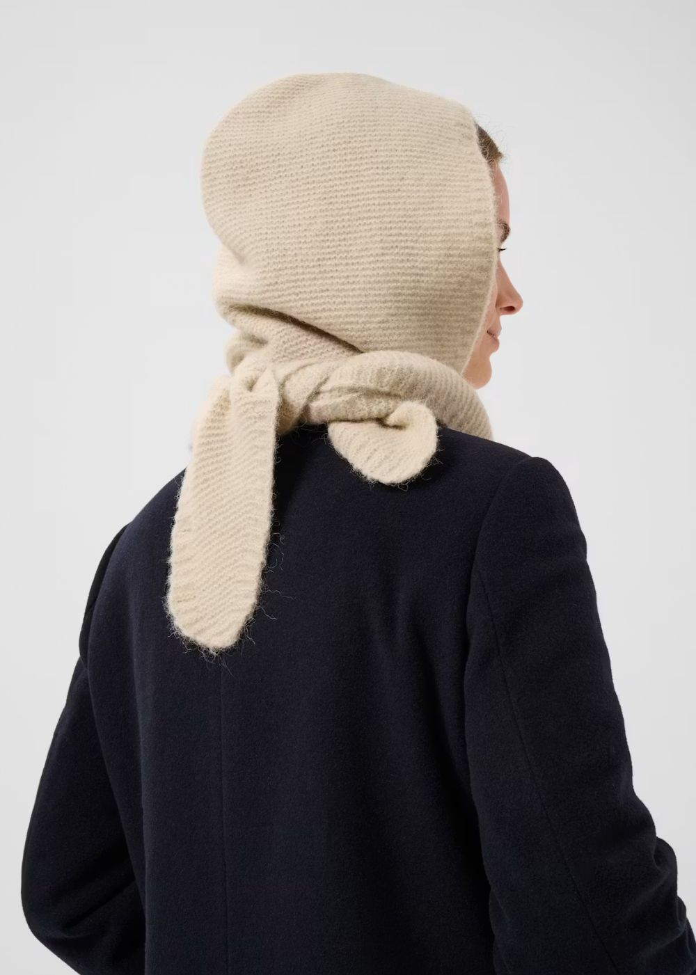 A model shown from the back wearing a cream-colored knit bonnet with long floppy bunny ears, paired with a dark navy coat against a light gray background.