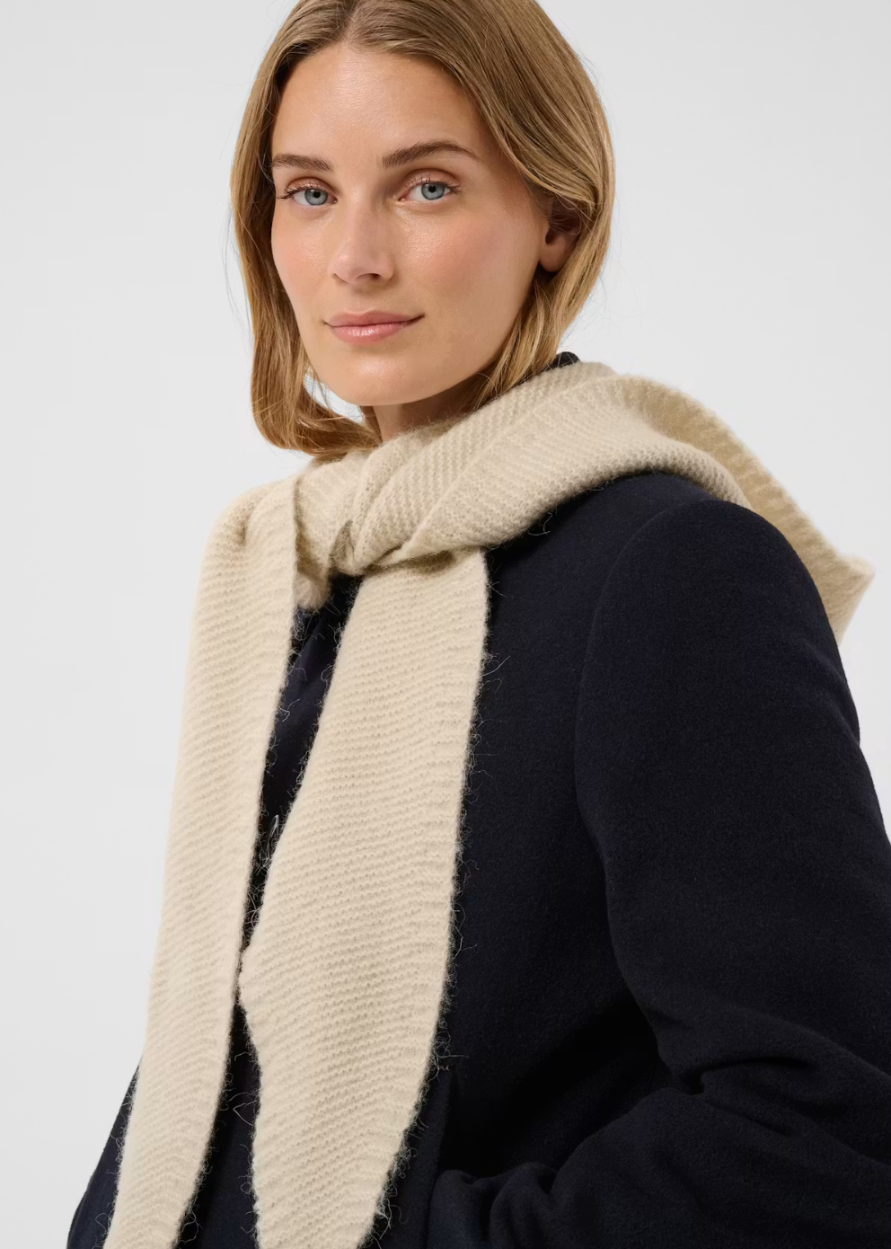 A woman wearing a thick, textured cream-colored knit scarf wrapped around her neck, paired with a dark navy coat, against a plain light gray studio background.
