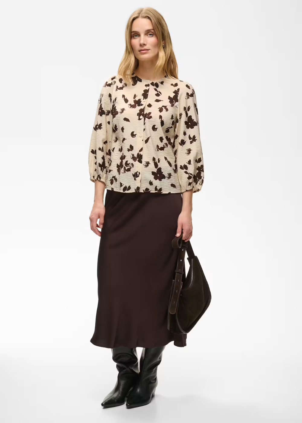 A model stands against a light gray studio background wearing a beige blouse with a dark brown floral pattern, a dark brown midi skirt, black pointed-toe boots, and holding a dark brown shoulder bag.