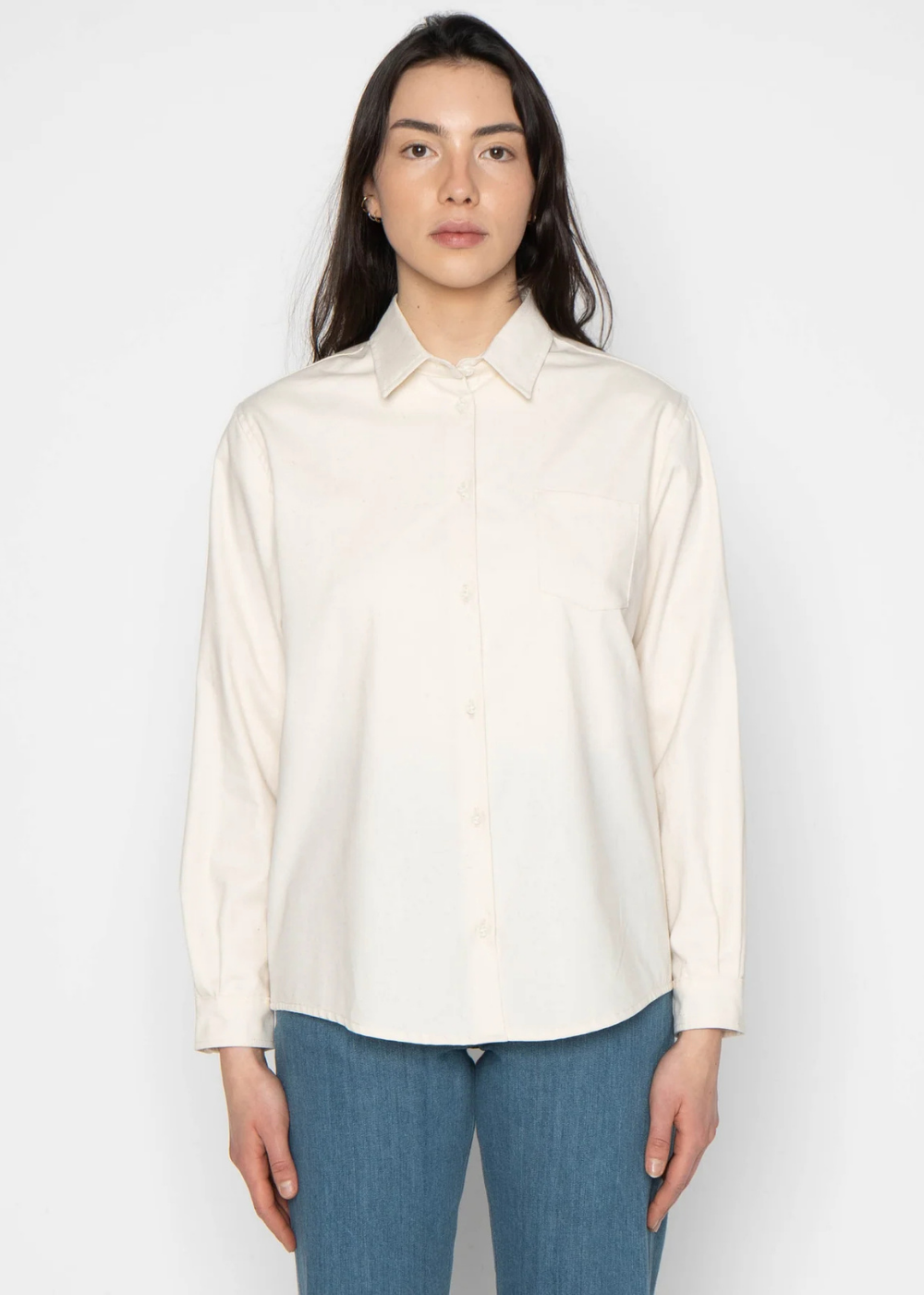 A woman modeling a long-sleeve cream-colored button-up shirt with a pointed collar, paired with blue jeans, against a plain light gray background.