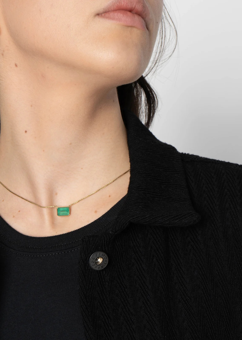 Close-up of a model wearing a delicate gold chain necklace with a small rectangular green pendant, paired with a black collared shirt.