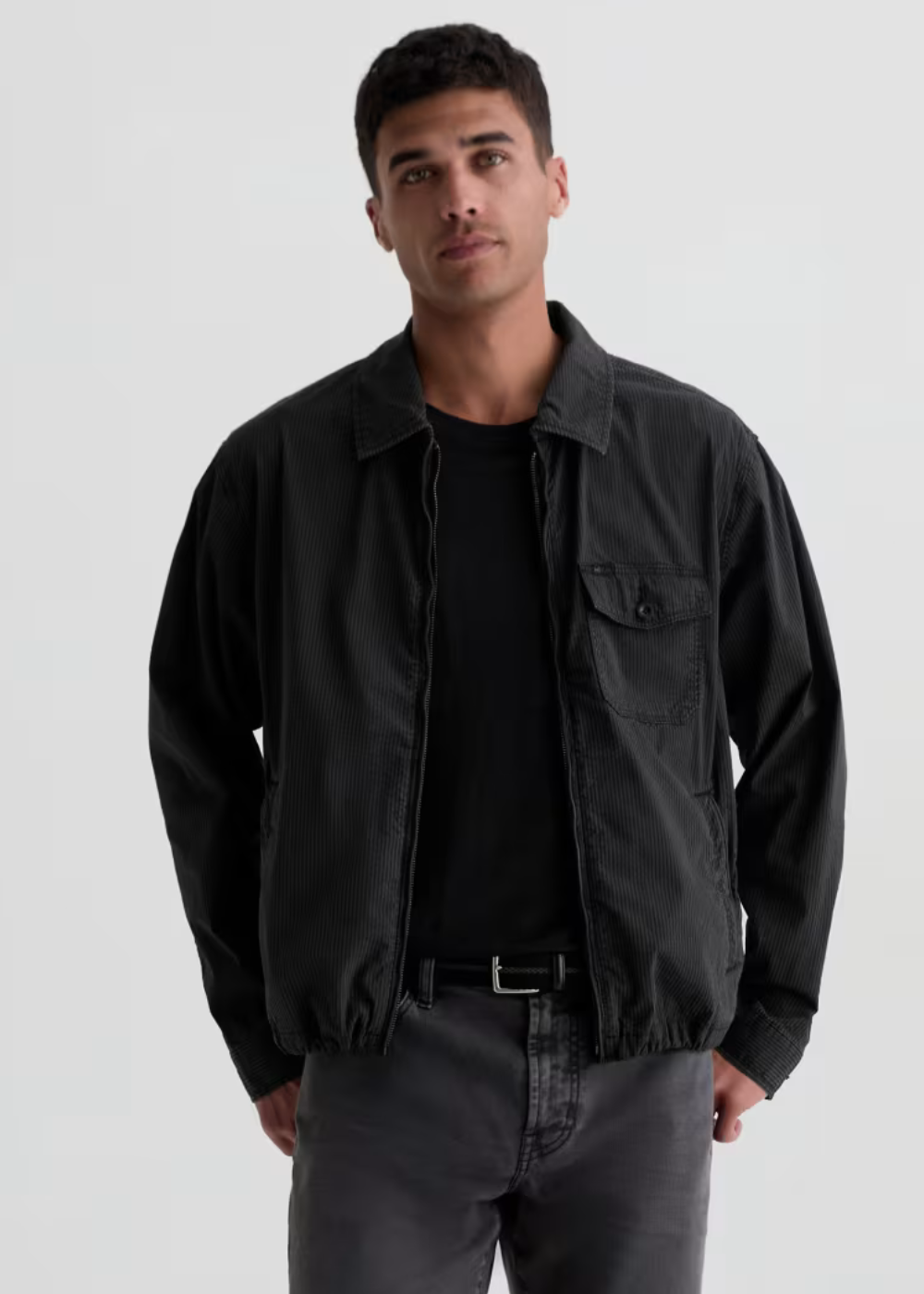 A man wearing a black lightweight jacket over a black t-shirt and dark gray jeans, standing against a light gray studio background.