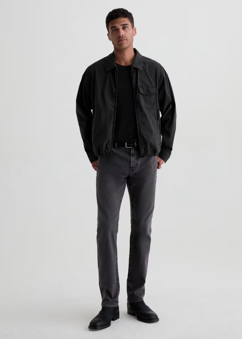A man modeling a black overshirt jacket over a black t-shirt, paired with dark gray trousers and black shoes, standing against a light gray studio background.