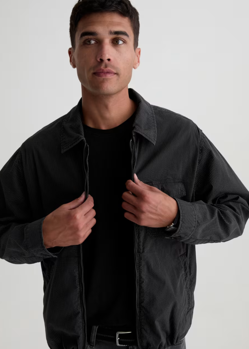 A man wearing a dark charcoal gray corduroy utility jacket over a black t-shirt, posing against a light gray studio background.