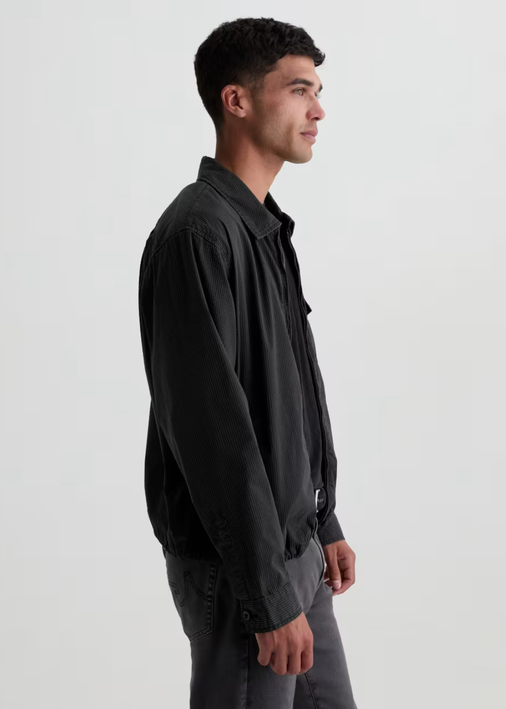 A man in side profile wearing a black button-up shirt jacket over a black shirt and gray trousers, photographed against a plain light gray studio background.