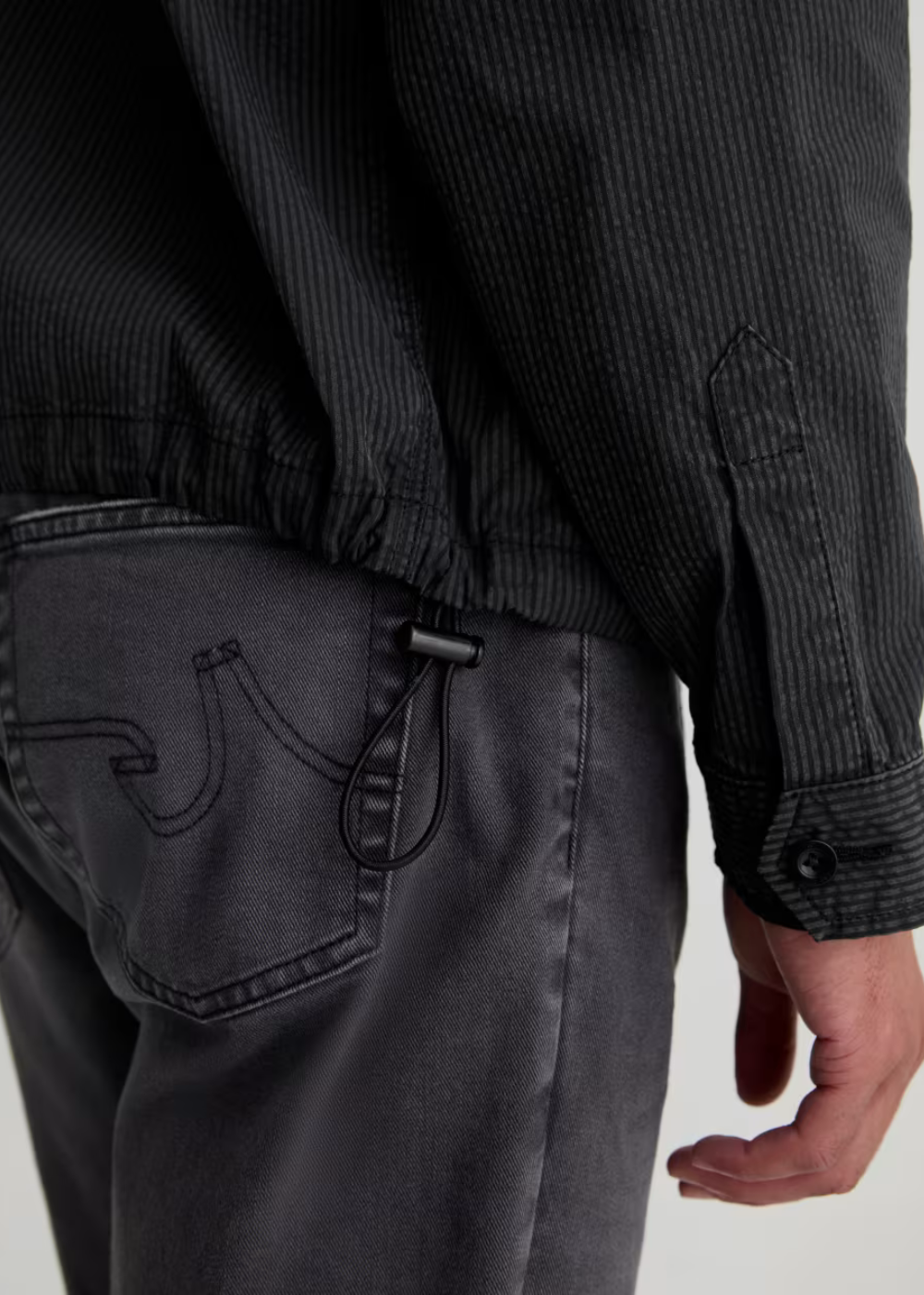 Close-up of a person wearing a dark charcoal corduroy jacket with an elasticated hem and a dark gray washed denim jacket with a distinctive curved pocket design.